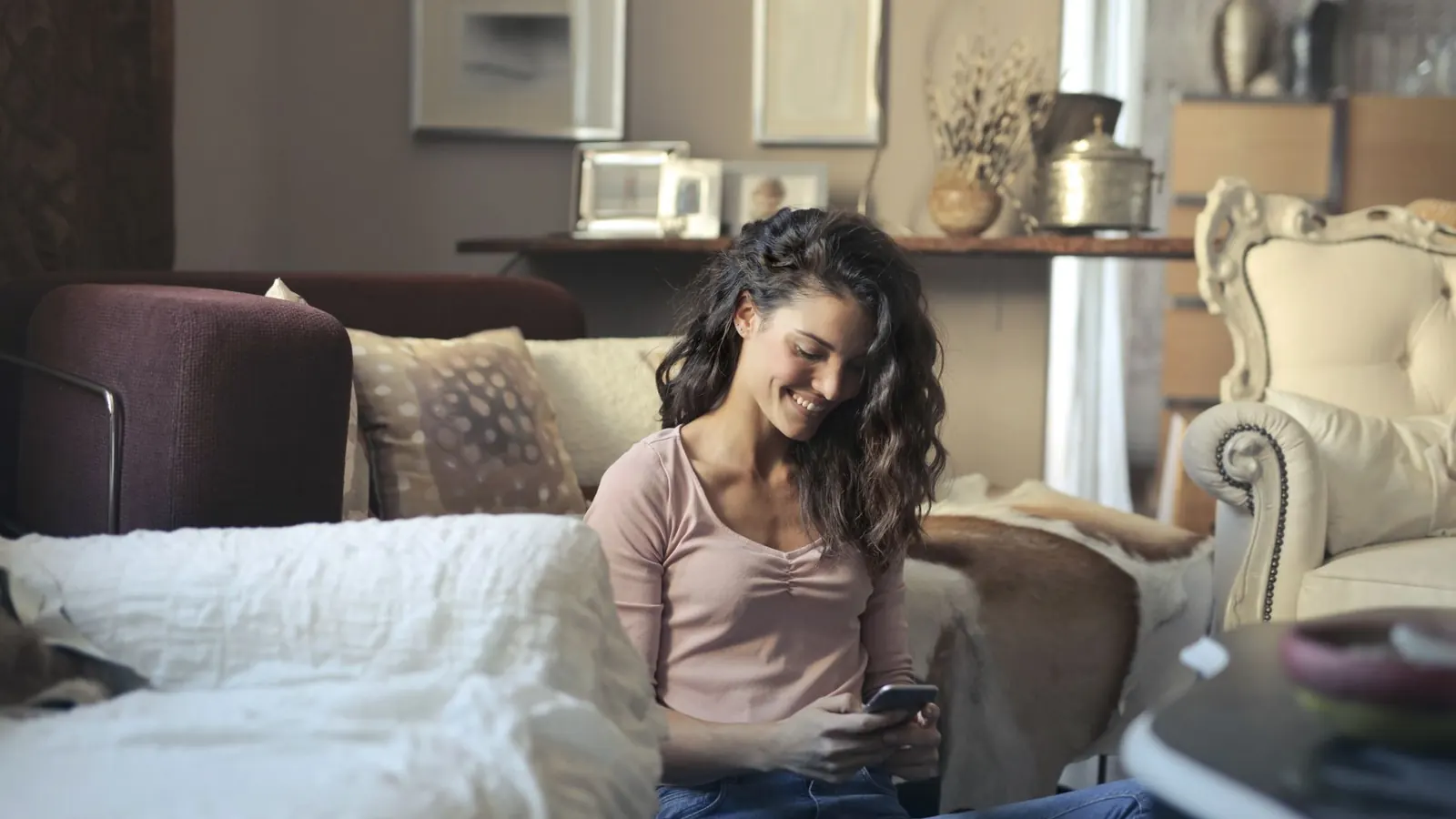 Mulher sorrindo sentada na sala de casa enquanto usa o celular