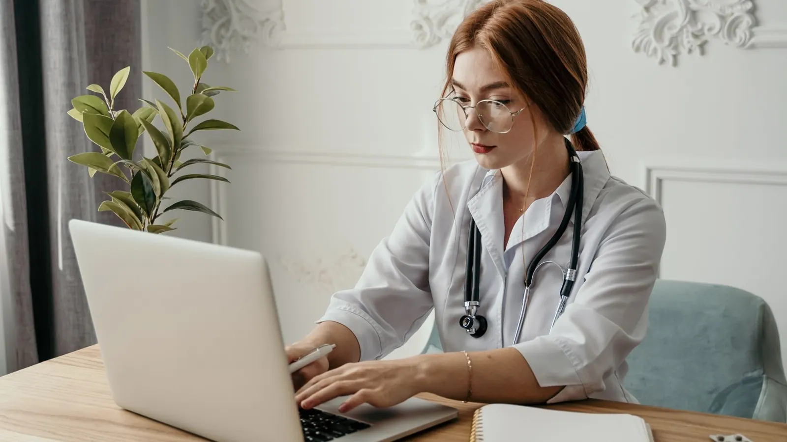 Médica de jaleco e estetoscópio digitando em um notebook na mesa do consultório