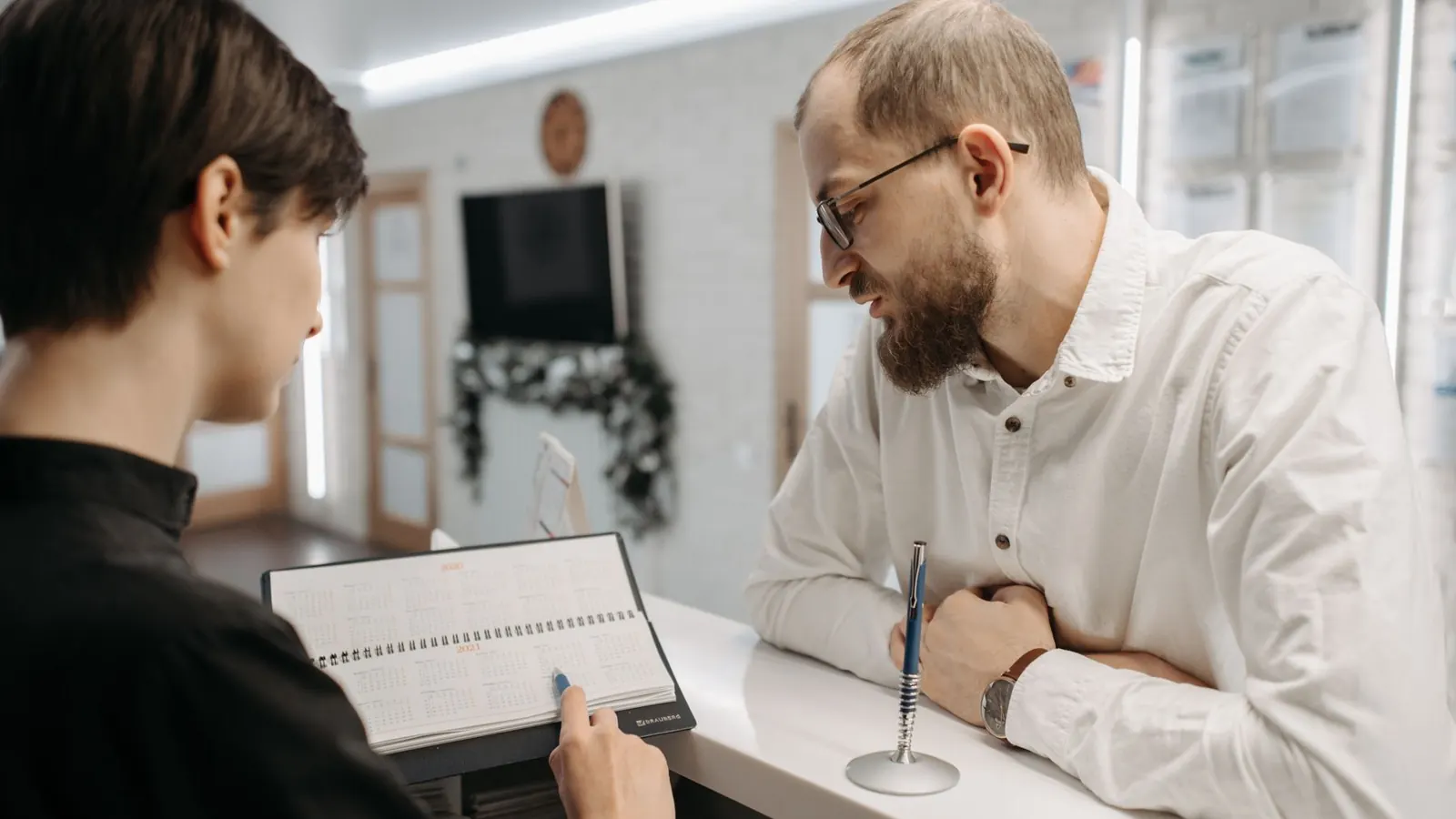 Recepcionista de clínica mostrando a agenda de papel a um paciente no balcão