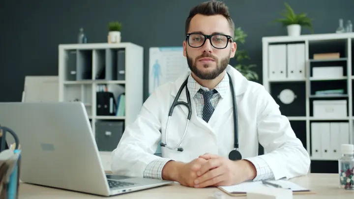 Médico de jaleco e óculos sentado à mesa do consultório com um notebook aberto