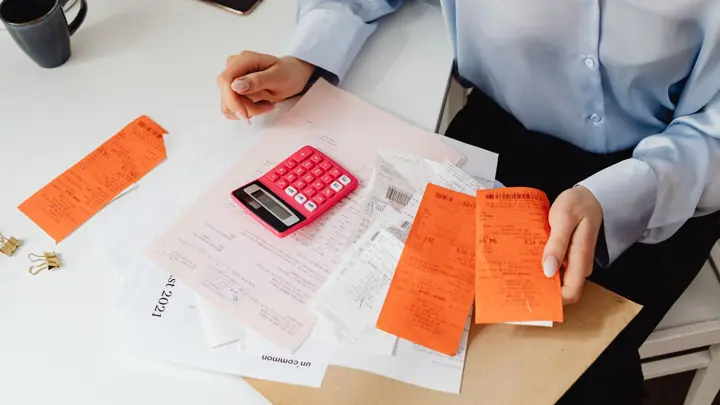 Mãos organizando recibos e notas ao lado de uma calculadora rosa sobre a mesa