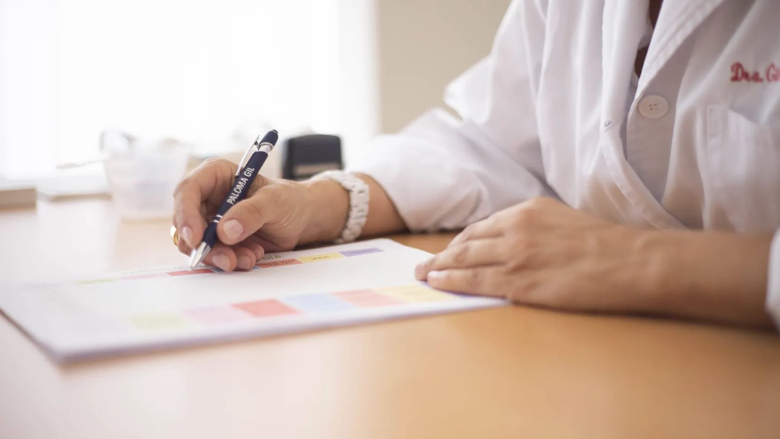 Mão de médica de jaleco escrevendo com caneta sobre um papel em cima da mesa do consultório